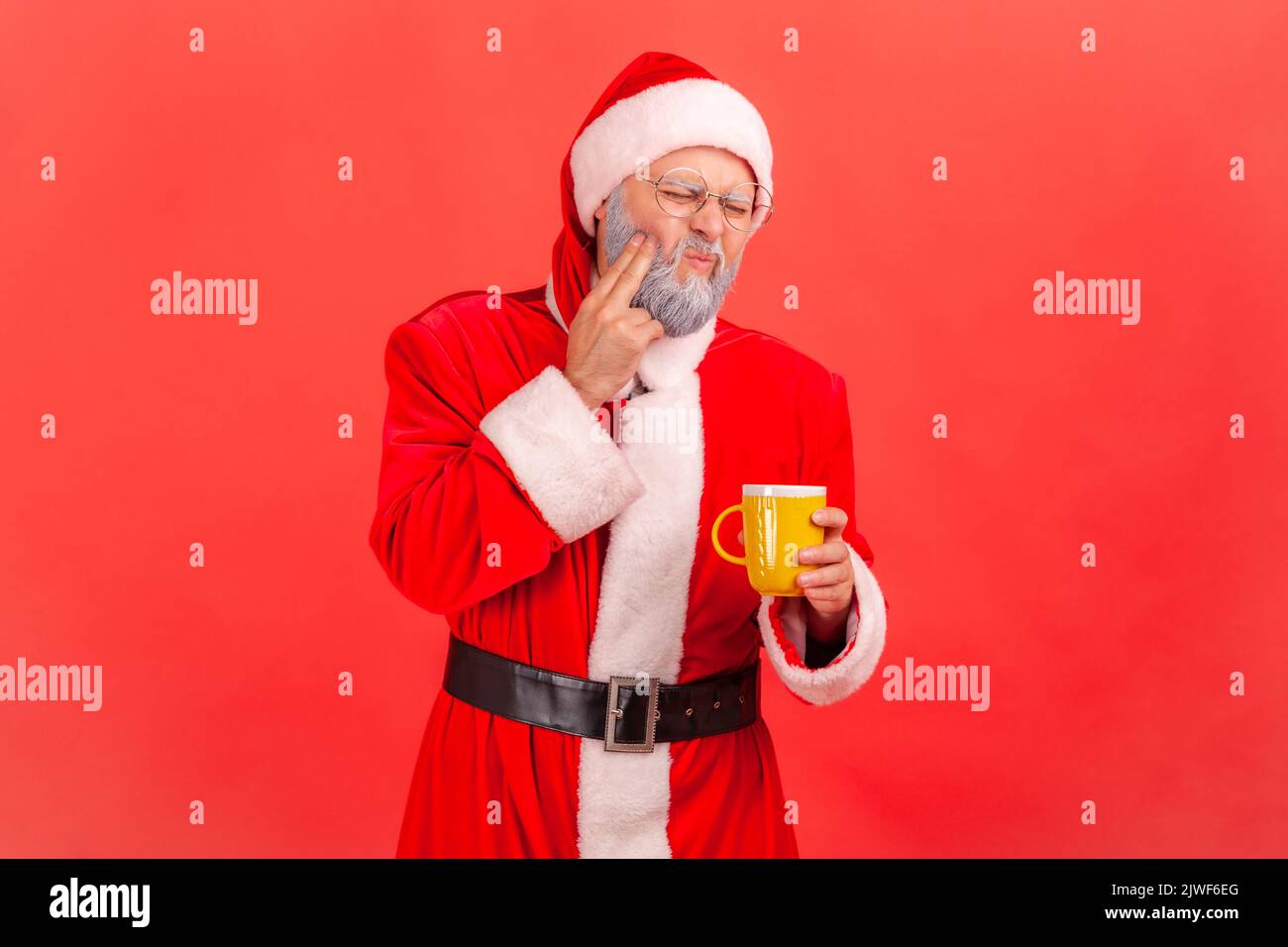 Ritratto dell'uomo anziano con barba grigia che indossa il costume di babbo natale che soffre di tremendo dolore ai denti dopo aver bevuto bevande calde o fredde. Studio in interni isolato su sfondo rosso. Foto Stock