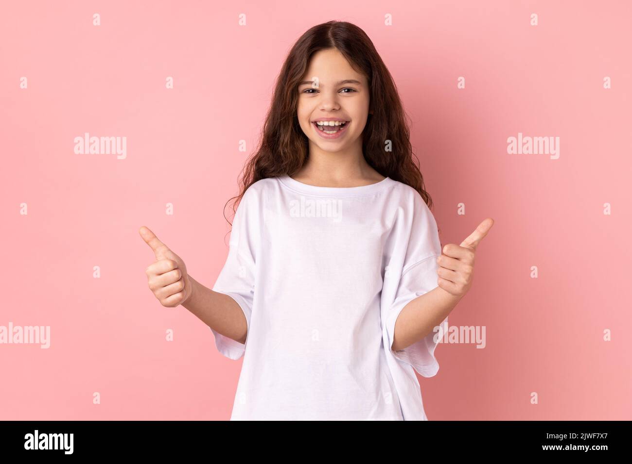 Ritratto di una bambina sorridente che indossa una T-shirt bianca che mostra i pollici, come un gesto, guardando la macchina fotografica dimostrando la sua soddisfazione. Studio in interni isolato su sfondo rosa. Foto Stock