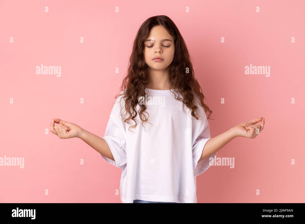 Ritratto di bambina rilassata che indossa una T-shirt bianca che fa yoga, respira, tiene gli occhi chiusi, calmando giù dopo dure lezioni. Studio in interni isolato su sfondo rosa. Foto Stock