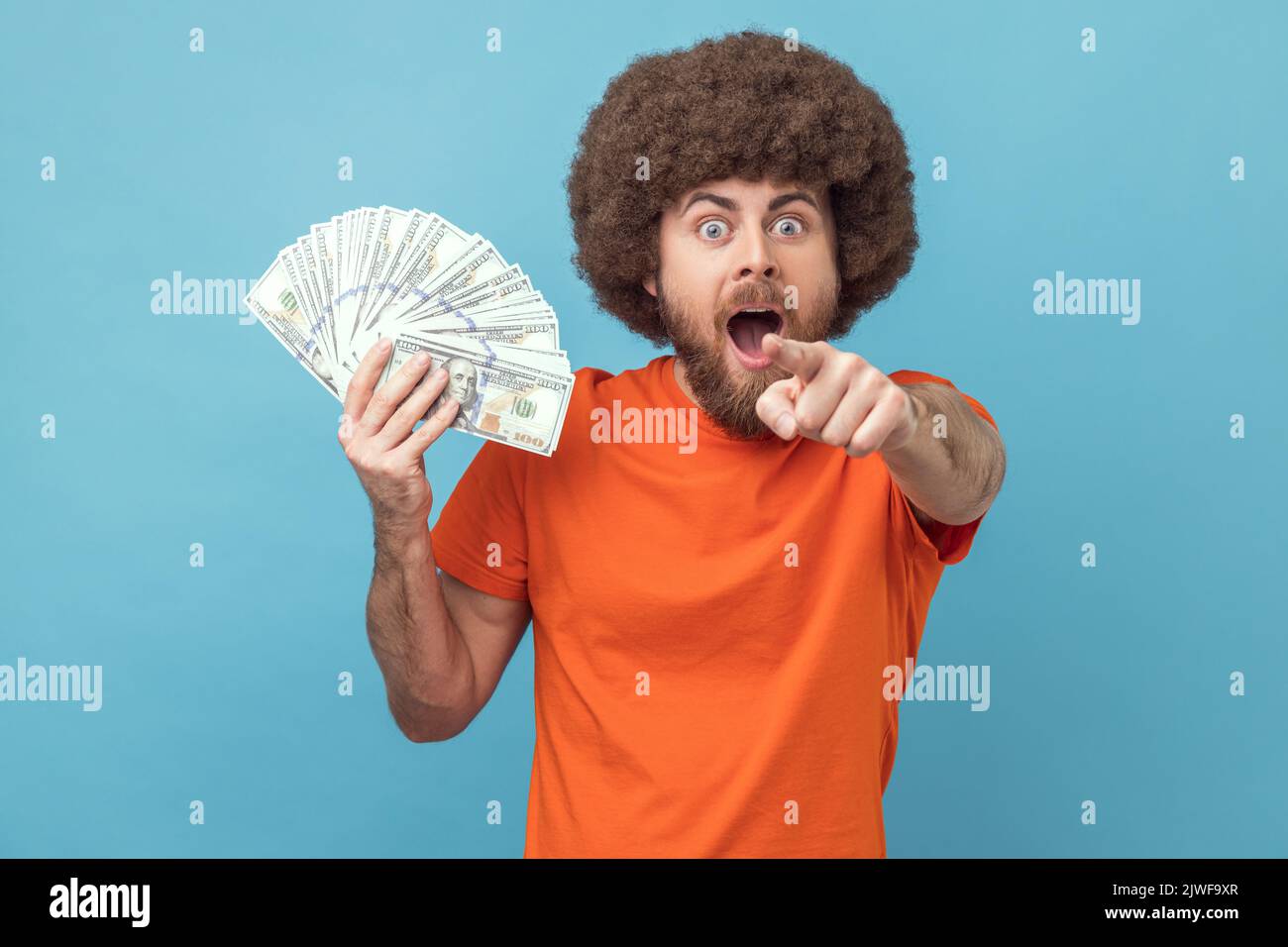 Ritratto di stupito scioccato uomo con acconciatura afro indossare T-shirt arancione in possesso di banconote in dollari, puntando alla fotocamera, si vince la lotteria. Studio in interni isolato su sfondo blu. Foto Stock