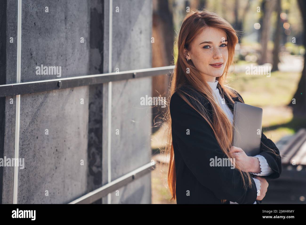 Bella donna d'affari in piedi con un computer portatile all'aperto nel parco cittadino, in attesa che la sua amica vada al lavoro. Allegra freelancer femminile . Lavoro in ufficio Foto Stock
