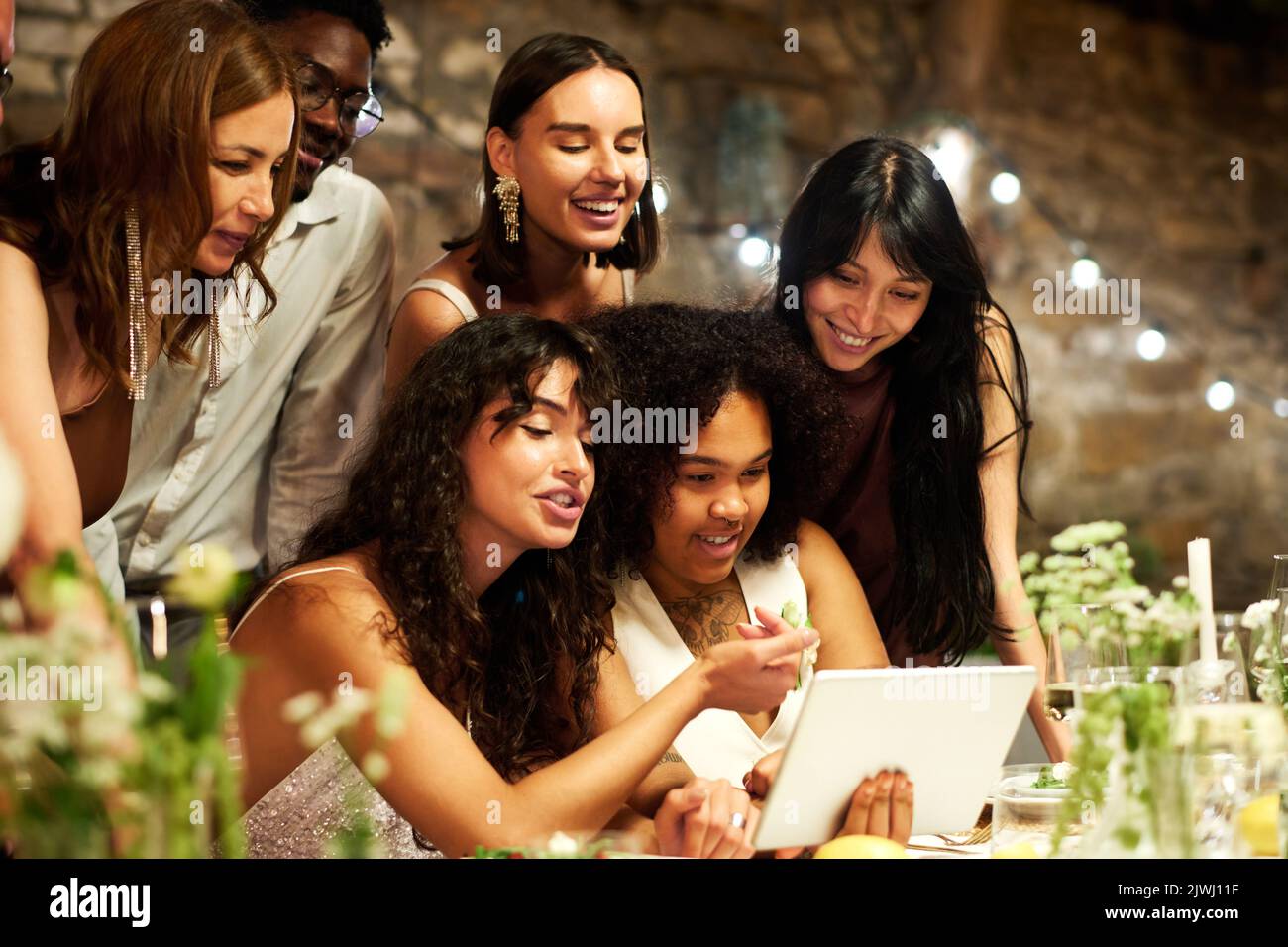 Due giovani spose interculturali e i loro amici comunicano in videochat mentre sono seduti al tavolo festivo servito durante la festa di nozze Foto Stock