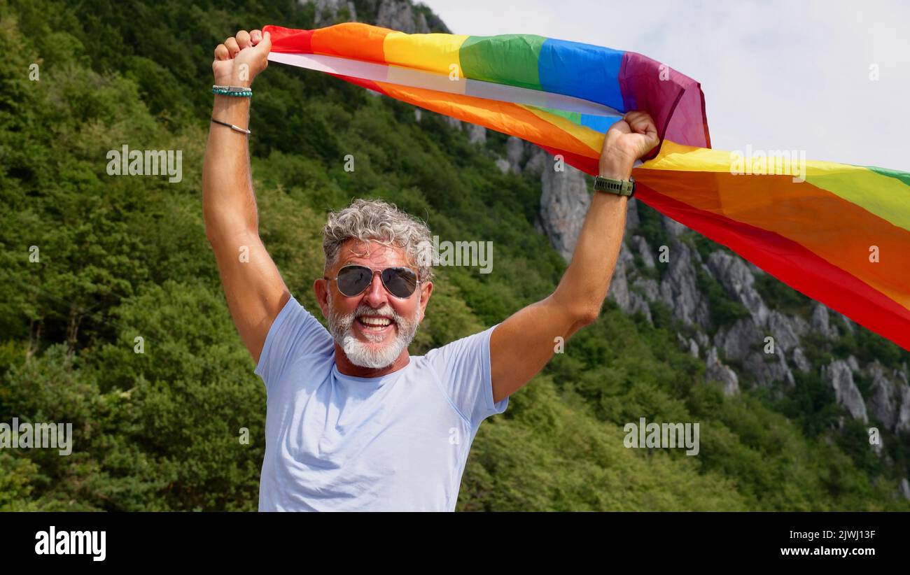 Ritratto di un anziano anziano dai capelli grigi bisessualità dell'uomo caucasico con barba e occhiali da sole in possesso di una bandiera arcobaleno LGBTQIA sulla natura. Celebra il mese dell'orgoglio, il giorno della bandiera dell'arcobaleno, la parata gay Foto Stock