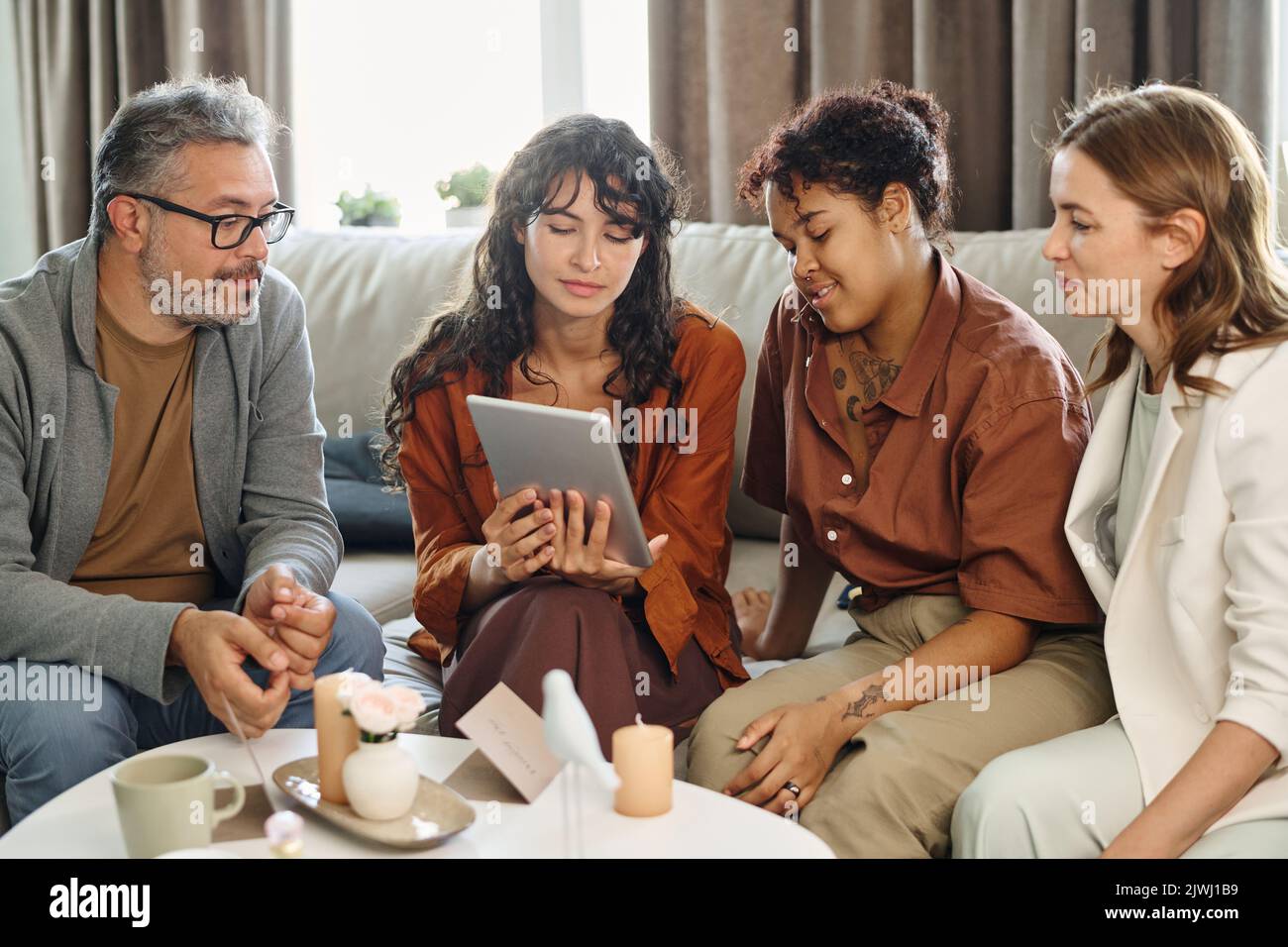 Giovane coppia lesbica interculturale in casualwear che mostra i genitori abiti da sposa sullo schermo del tablet tenuto dalla loro figlia Foto Stock