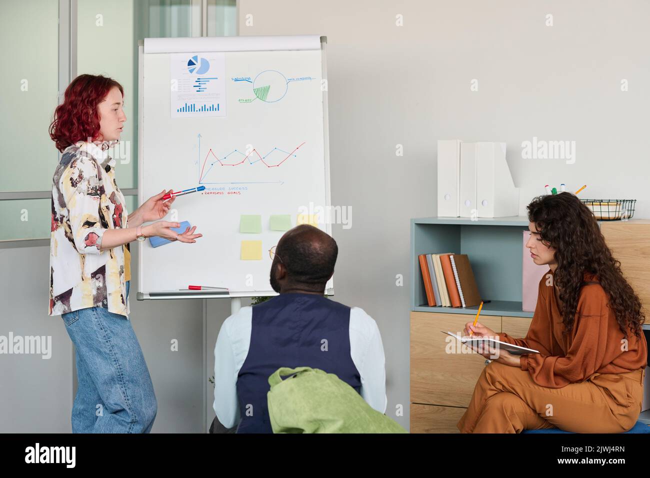 Giovane broker fiducioso in casualwear che punta a whiteboard con grafico finanziario mentre spiega le fluttuazioni delle finanze Foto Stock