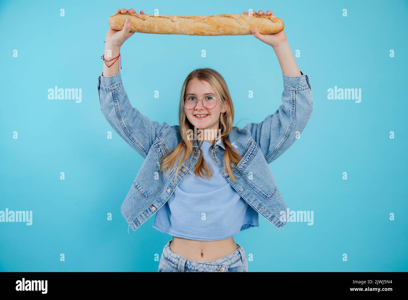 Ragazza adolescente sottile tenere sopra la testa gustosa baguette francese su sfondo blu vuoto, spazio copia libero. Foto di giovane donna sorridente e alla moda che mostra fresco Foto Stock