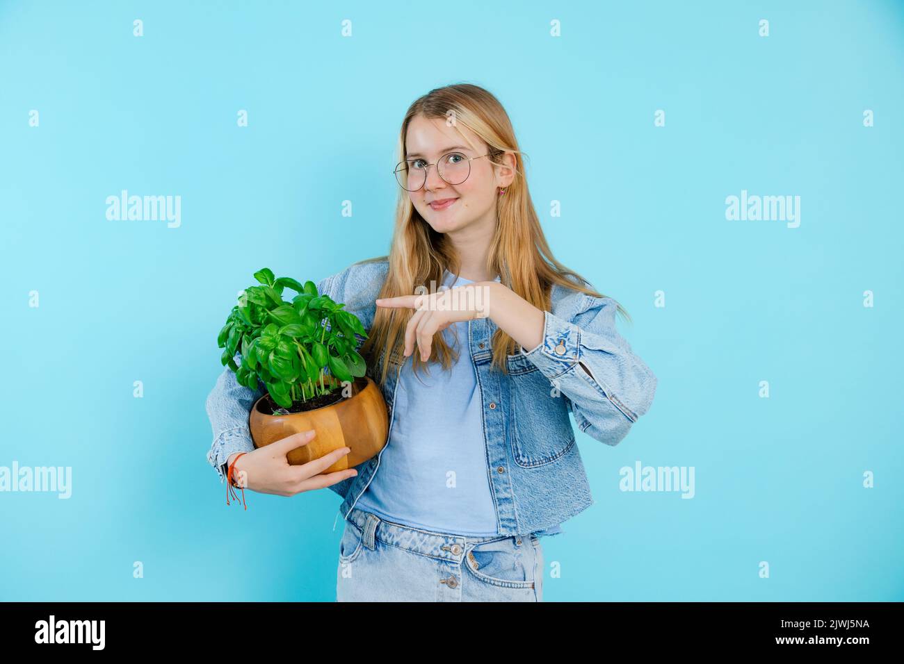 Felice ragazza teenage mostra e punto a pianta di casa verde in pentola di legno su sfondo blu vuoto, spazio di copia gratuito. Foto di giovane donna sorridente in crescita Foto Stock