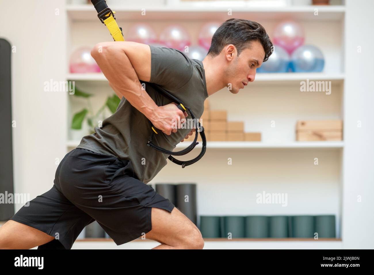 Vista laterale dell'uomo in forma concentrata che si esercita sui cinturini TRX sospesi mentre si allena nel moderno e spazioso centro fitness Foto Stock