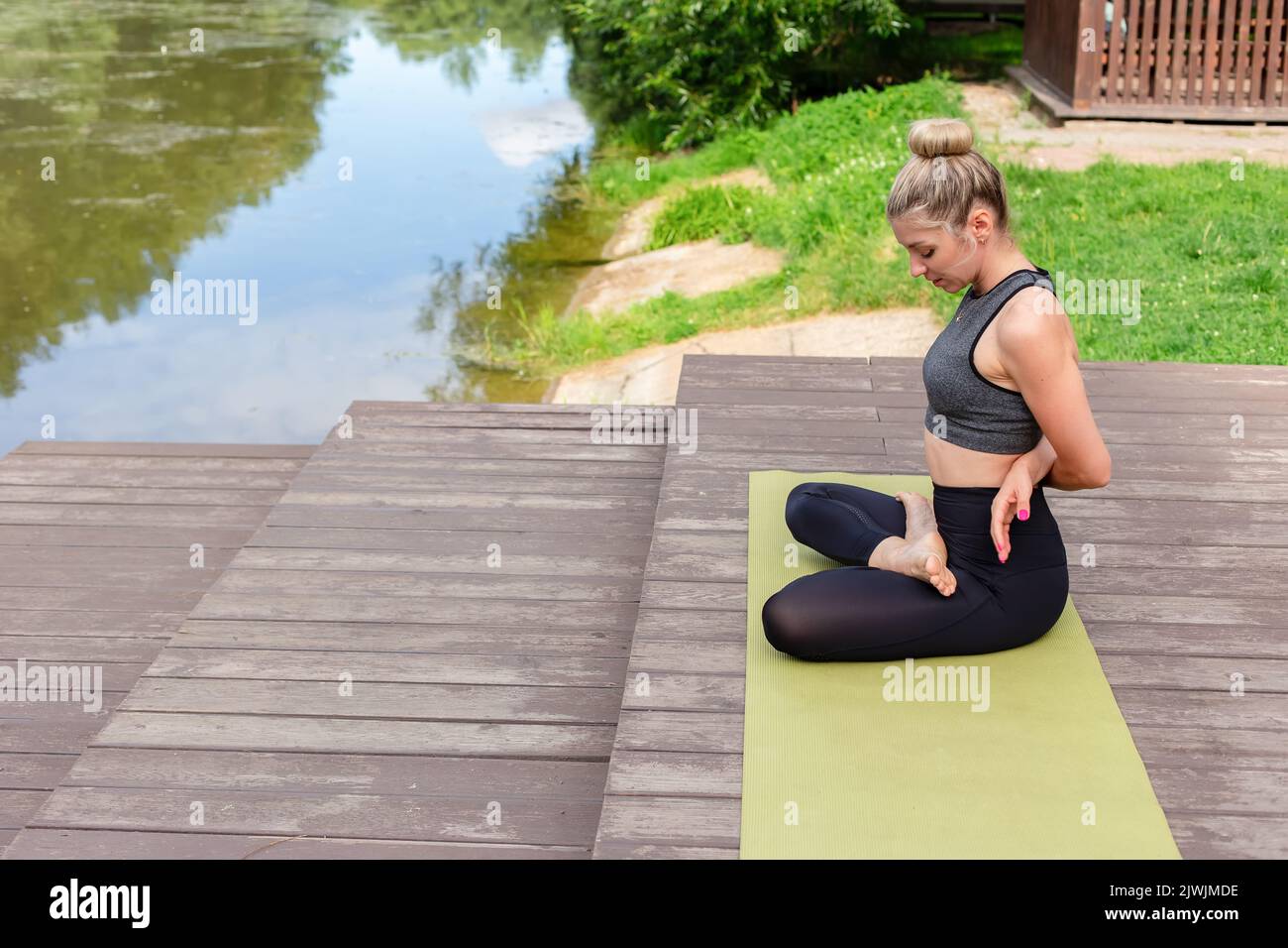 Una donna snella seduta su una piattaforma di legno vicino ad uno stagno nel parco in estate e che esegue yoga Foto Stock