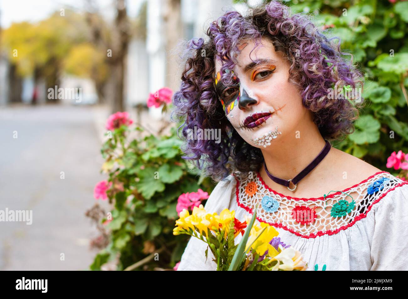 giovane argentina latina con la Calavera Catrina trucco e abiti folcloristici guardando la macchina fotografica con un volto pensivo e tenendo fiori è s Foto Stock