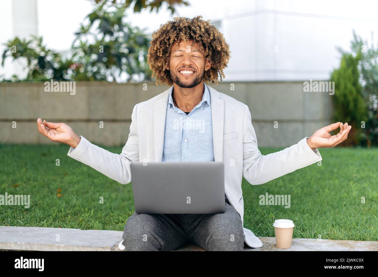 Pausa durante il lavoro. Calma rilassato ricci capelli ispanici o brasiliano uomo, ufficio dipendente, si siede all'aperto vicino al business center, medita, si rilassa, chiuso gli occhi, si prende cura della salute mentale, sorriso Foto Stock