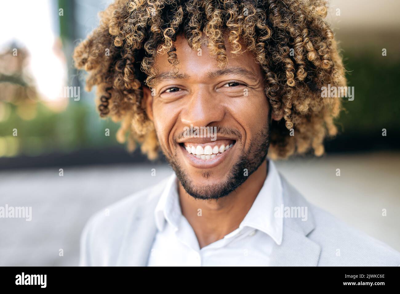 Foto ravvicinata di un bel latino o un uomo brasiliano dai capelli ricci, elegante, elegante e di successo nelle gare miste, in piedi all'aperto, guardando la fotocamera, sorridente Foto Stock