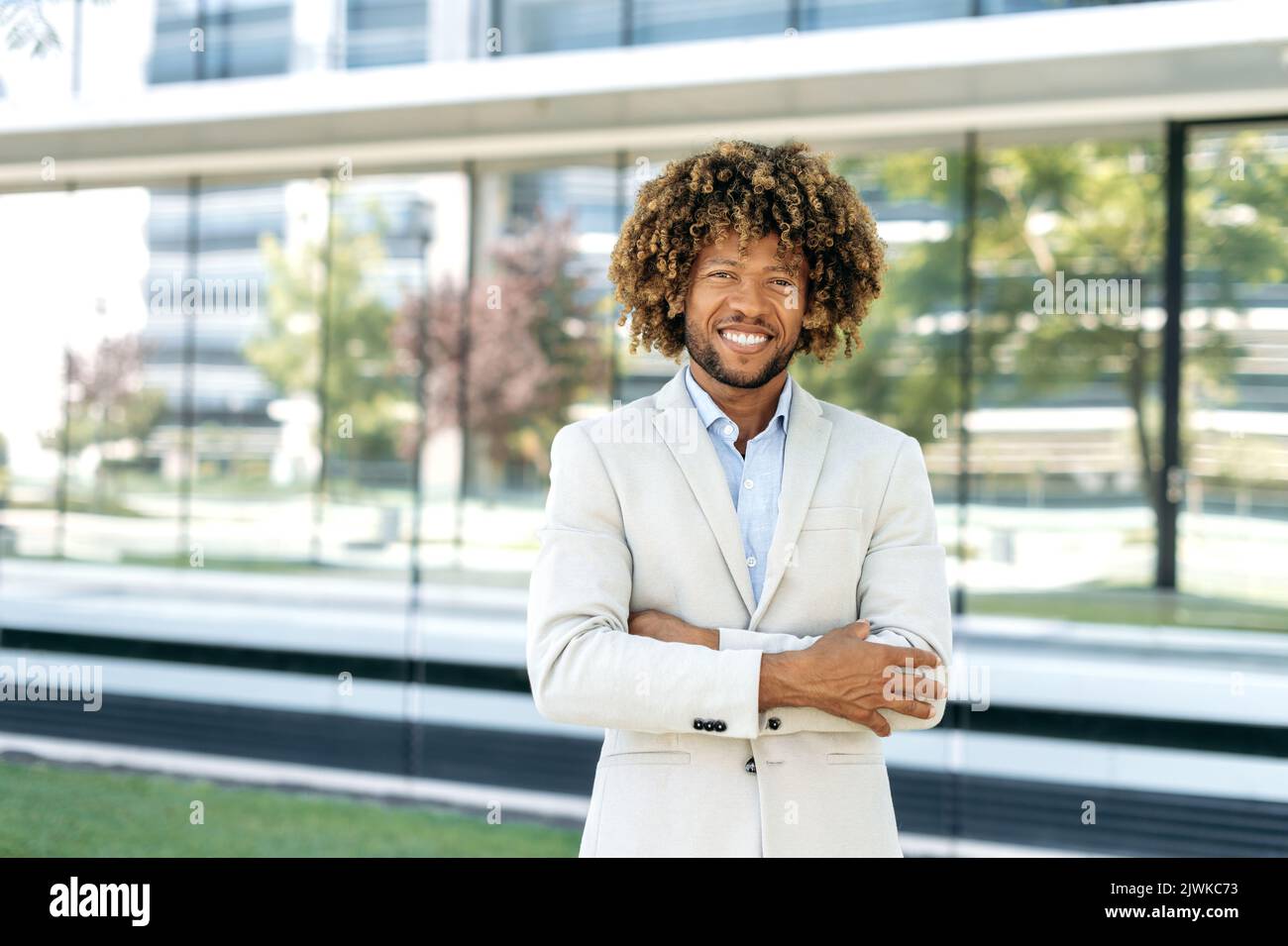 Ritratto di un uomo di razza, imprenditore, dirigente aziendale, elegante e di successo, riccio misto, in piedi all'aperto sullo sfondo di un business center con braccia incrociate, guardando la macchina fotografica, sorridendo Foto Stock