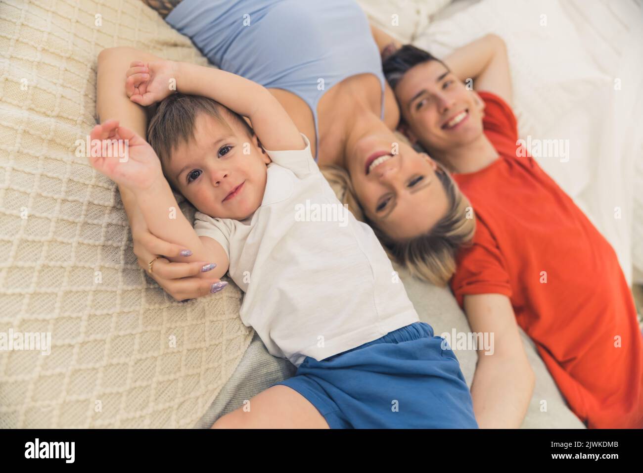 Vista dall'alto di una famiglia di tre persone - il ragazzo caucasico in maglietta bianca, la sua bella madre bionda in canotta blu chiaro e il suo padre sorridente in maglietta arancione - sdraiato sul letto e divertendosi. Foto di alta qualità Foto Stock