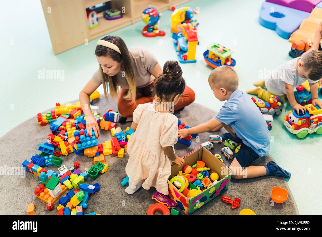 I bambini e il loro insegnante di asilo giocano con blocchi di plastica e giocattoli colorati per auto mentre si siedono sul pavimento in una sala giochi. Concentrazione, sviluppo delle capacità motorie fini e lorde, . Foto di alta qualità Foto Stock