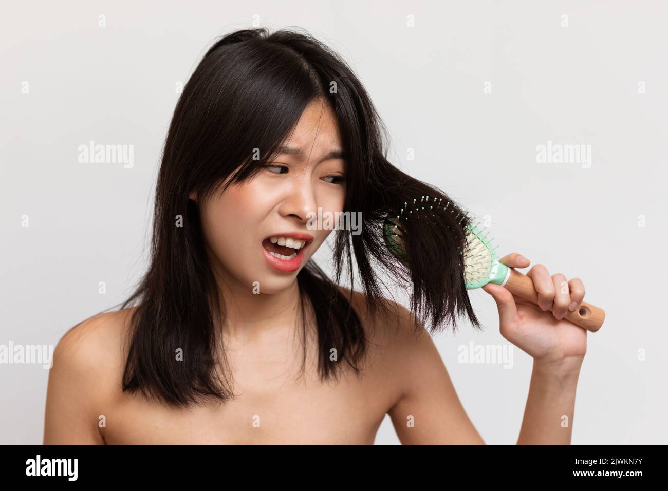 Cosmetici per capelli secchi e fragili. La signora coreana irritata non può pettinare i capelli, in piedi su sfondo bianco studio Foto Stock