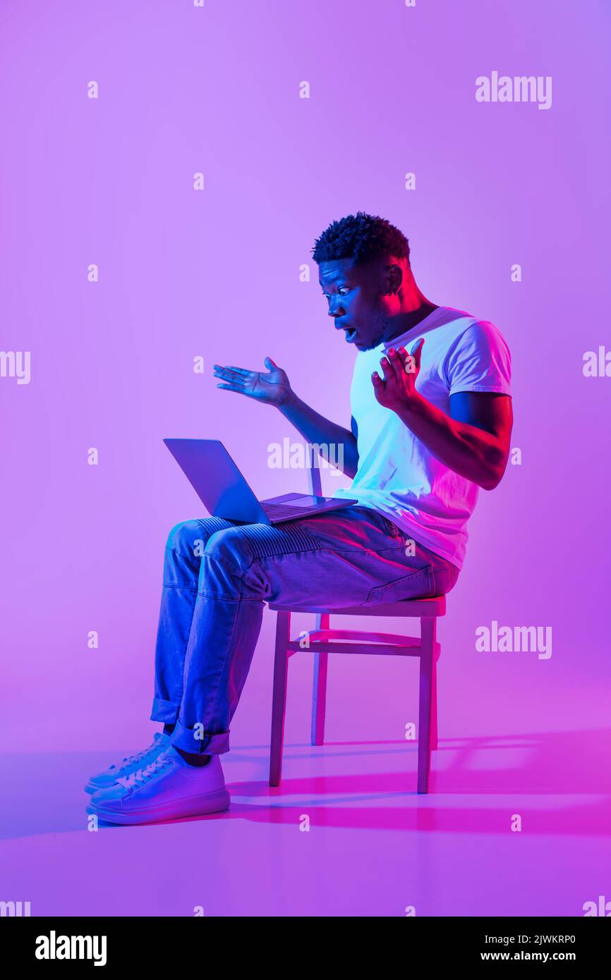 Emotivo giovane uomo nero seduto sulla sedia con notebook, sensazione di scioccata, facendo errore nel progetto di business in luce al neon Foto Stock