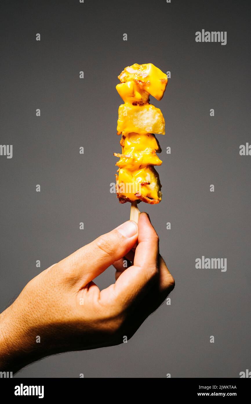 Mano umana che tiene una brocetta di patate fritte con formaggio fuso e sormontata con pancetta su uno sfondo grigio Foto Stock