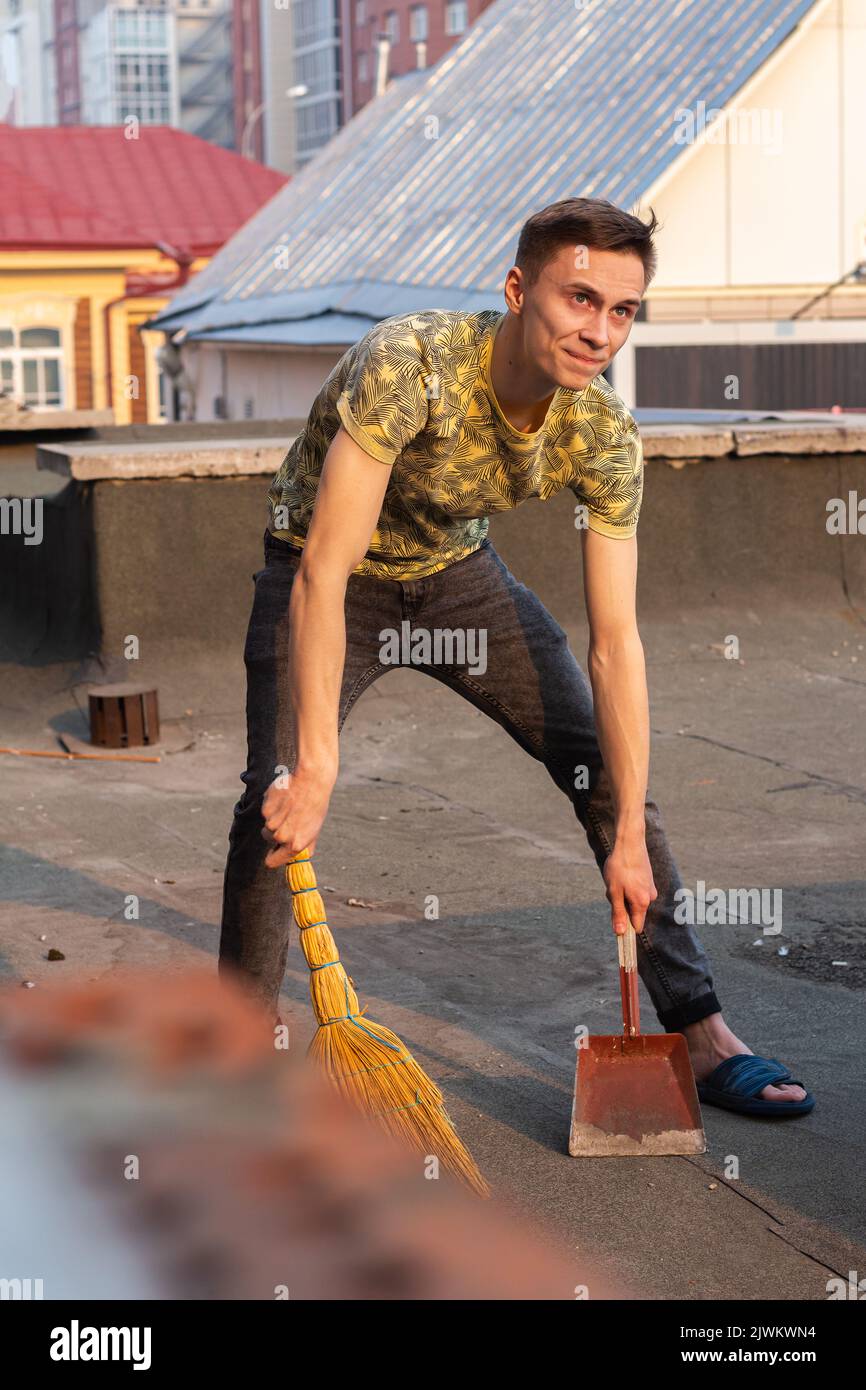 giovane uomo, ragazzo, pulisce con scopa sul tetto della sua casa, è arrabbiato con i vicini che buttano via la spazzatura. il concetto di pulizia, selettivo f Foto Stock