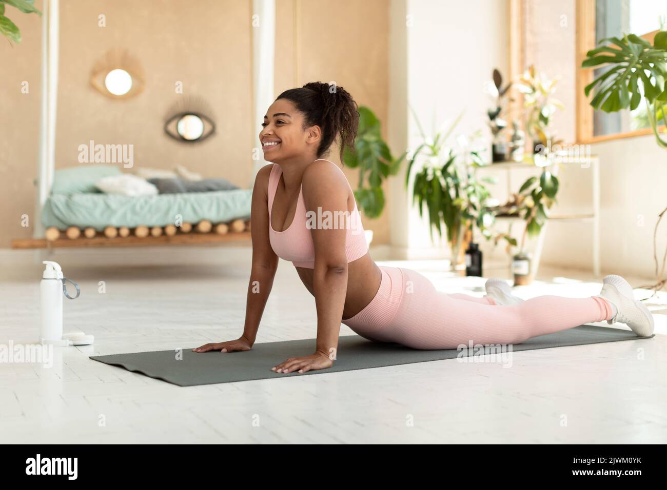 Felice vestito nero donna che fa yoga cobra posa, esercizio sul tappeto in camera da letto accogliente interno, copia spazio Foto Stock