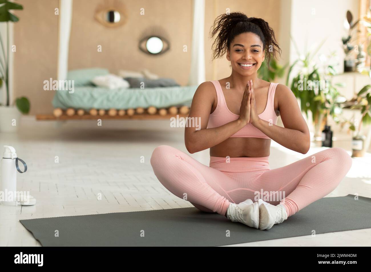 Concetto di yoga mattutino. Felice signora nera che pratica la meditazione in posizione lotus, seduta con le mani afferrate e sorridente Foto Stock