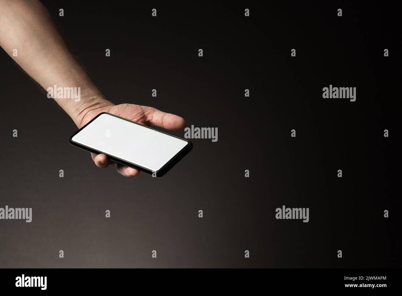 La mano dell'uomo ha teso lo smartphone nel palmo della mano con lo schermo bianco vuoto isolato su sfondo scuro con spazio di copia Foto Stock