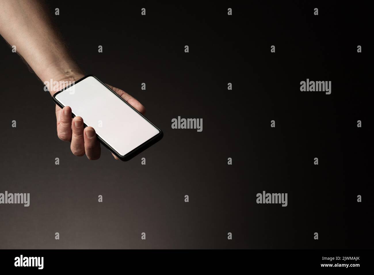 La mano dell'uomo ha teso lo smartphone nel palmo della mano con lo schermo bianco vuoto isolato su sfondo scuro con spazio di copia Foto Stock
