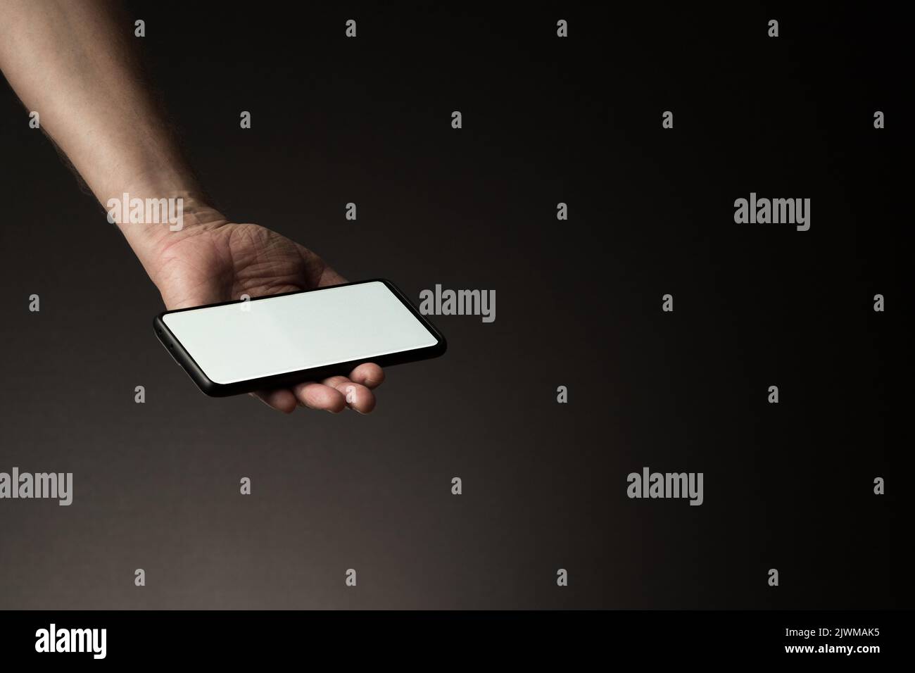 La mano dell'uomo ha teso lo smartphone nel palmo della mano con lo schermo bianco vuoto isolato su sfondo scuro con spazio di copia Foto Stock