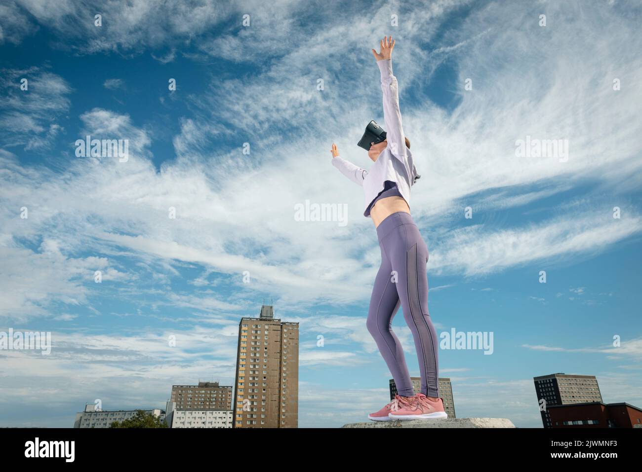 Donna all'aperto che indossa cuffie per la realtà virtuale con le braccia allungate e lo sfondo urbano. Foto Stock