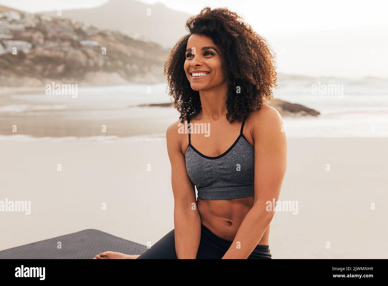 Ritratto di una bella giovane donna con capelli ricci indossando abbigliamento sportivo seduta sulla spiaggia rilassante al tramonto Foto Stock