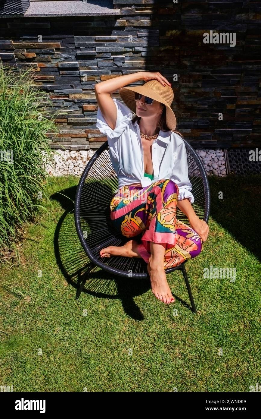 Scatto ad alta angolazione di una donna attraente che indossa una camicia bianca e un cappello da sole mentre si rilassa in una poltrona in giardino. Foto Stock