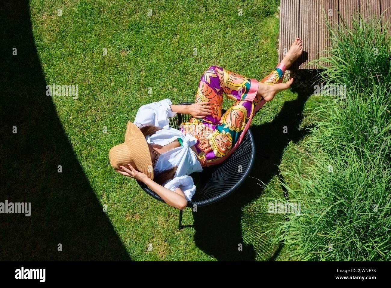 Scatto ad alta angolazione di una donna attraente che indossa una camicia bianca e un cappello da sole mentre si rilassa in una poltrona in giardino. Foto Stock