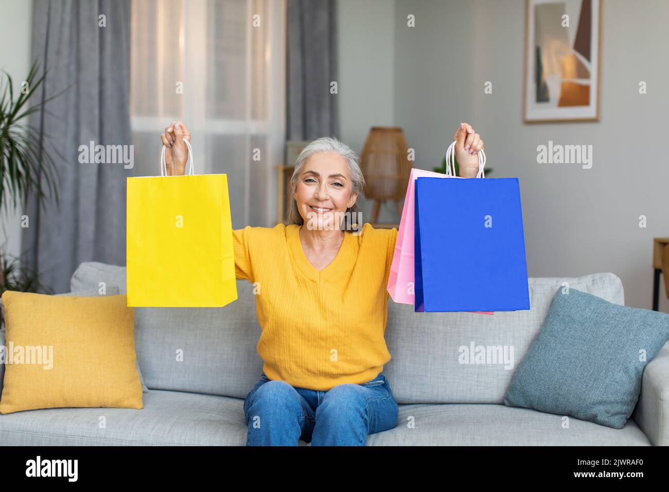 Allegro caucasico maturo grigio-capelli donna mostra pacchetti colorati con acquisti, si siede sul divano Foto Stock