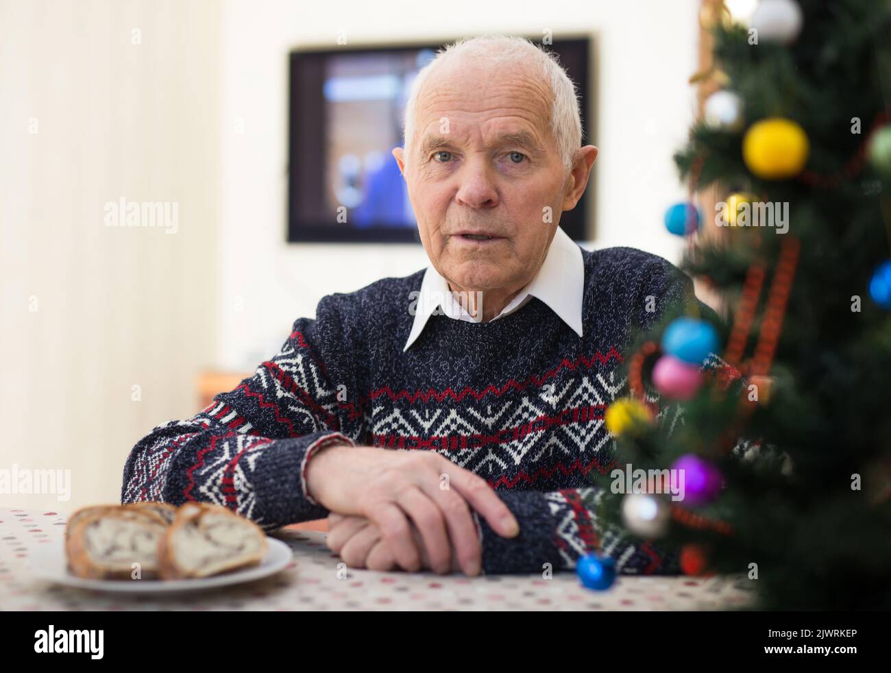 Uomo anziano positivo seduto da solo a casa con albero di Natale Foto Stock
