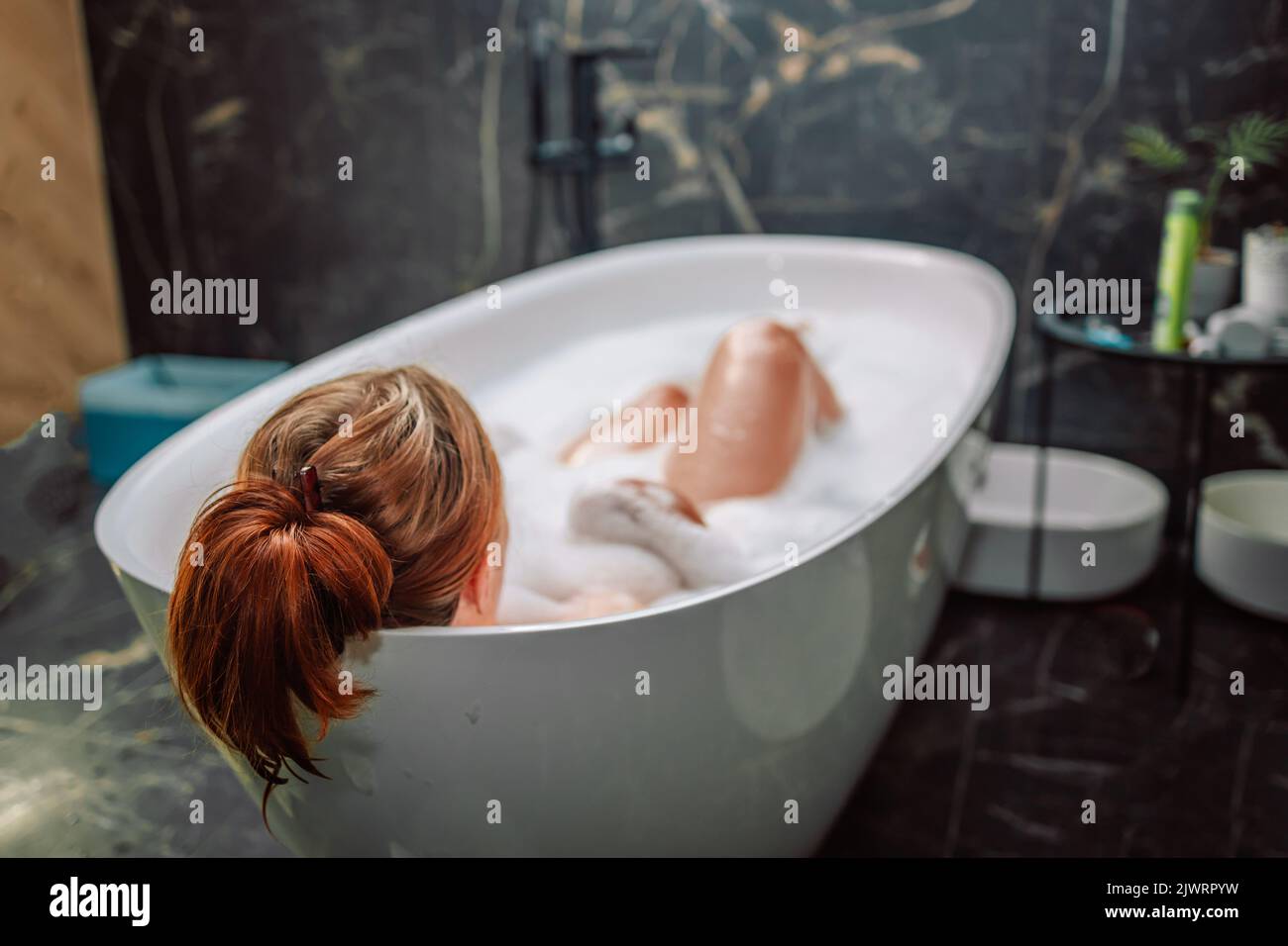 Vista posteriore di una giovane donna che ha il bagno di bolla e che guarda la finestra. Donne che si rilassano nella vasca da bagno. Foto Stock