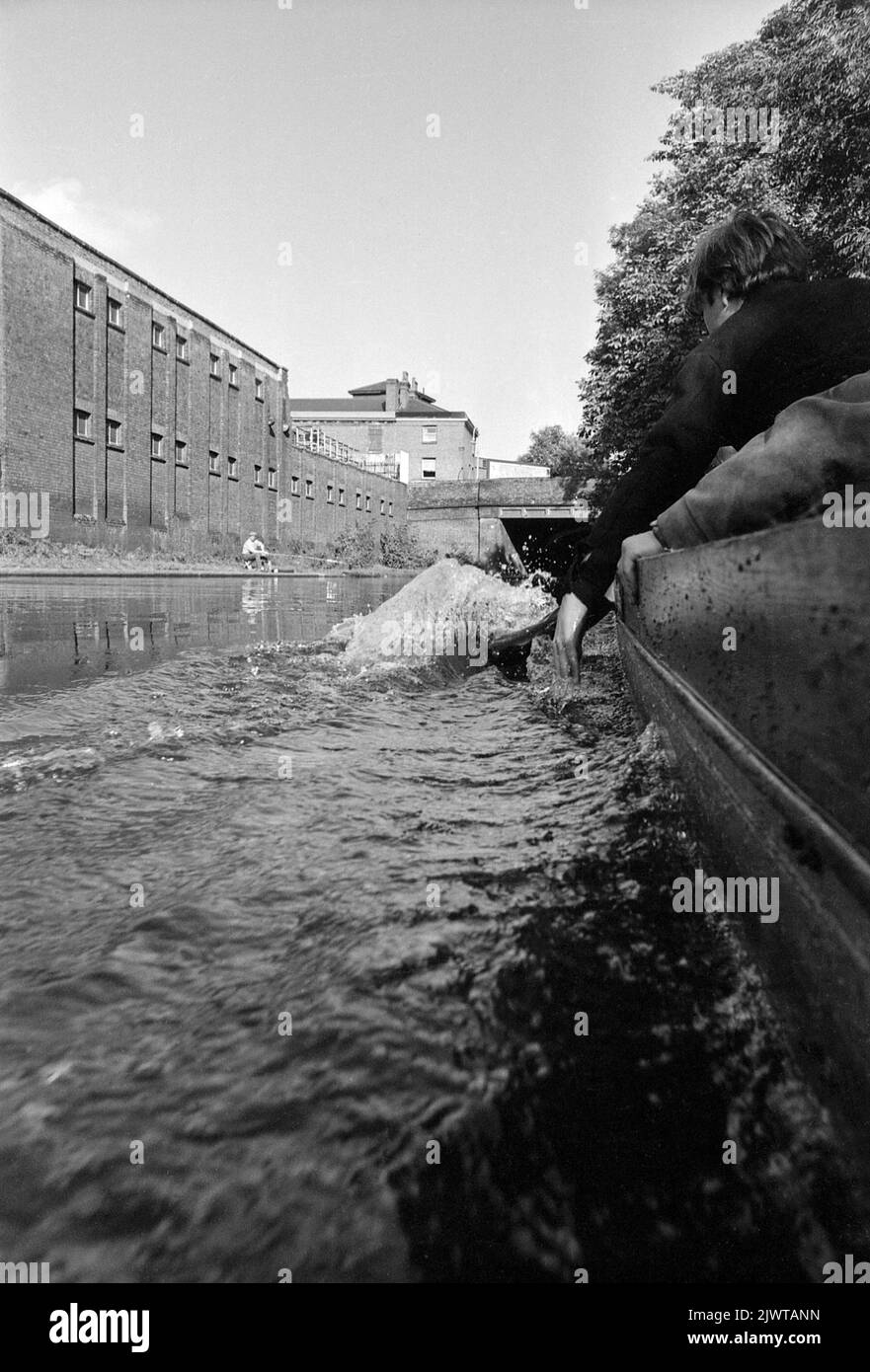 Londra, Inghilterra, circa 1967. Ragazzi del Pirate Club a bordo di una piccola barca sul Regent’s Canal. Un ragazzo sta trascinando le dita attraverso l'acqua. Un pescatore dal towpath guarda sopra. Il Pirate Club, un club nautico per bambini, è stato fondato nel 1966 presso Gilbey’s Wharf sul Regent’s Canal vicino a Camden, Londra. La loro clubhouse era una vecchia chiatta e un certo numero di piccole barche e canoe erano state donate per l’uso dei bambini. Foto Stock