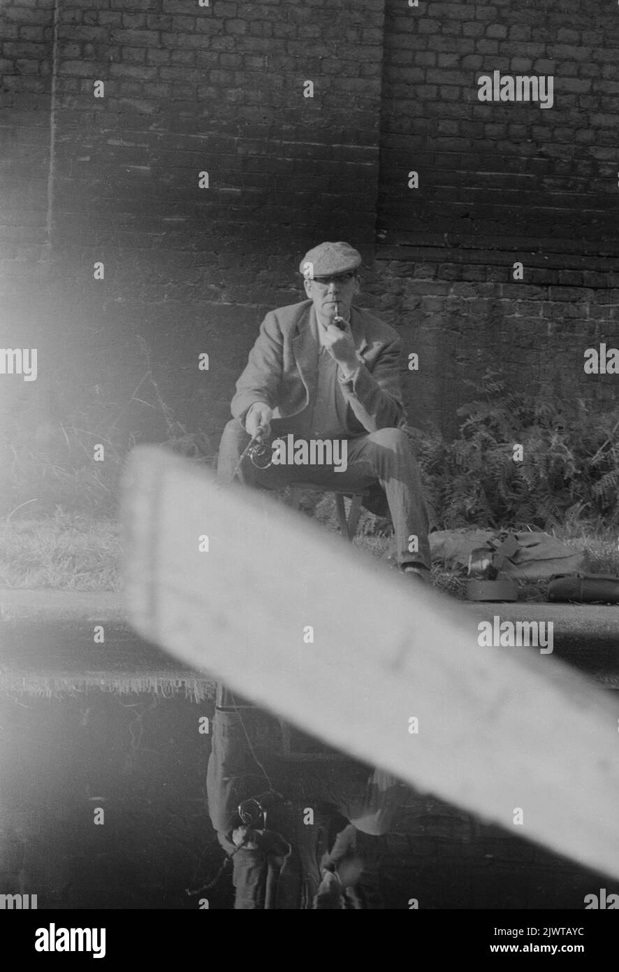 Londra, Inghilterra, circa 1967. Un pescatore, fumando una pipa, sta pescando dall'alzaia sul Regent's Canal a Camden tra Oval Road e Gloucester Road. La fotografia è stata scattata da una barca appartenente al Pirate Club. Un remo è visibile in primo piano. Il Pirate Club, un club nautico per bambini, è stato fondato nel 1966 presso Gilbey’s Wharf sul Regent’s Canal vicino a Camden, Londra. La loro clubhouse era una vecchia chiatta e un certo numero di piccole barche e canoe erano state donate per l’uso dei bambini. Foto Stock