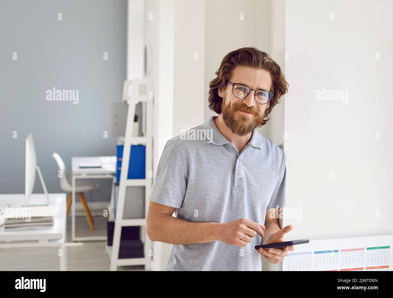 Uomo d'affari felice in T-shirt in piedi in ufficio, tenendo il telefono cellulare e sorridendo Foto Stock
