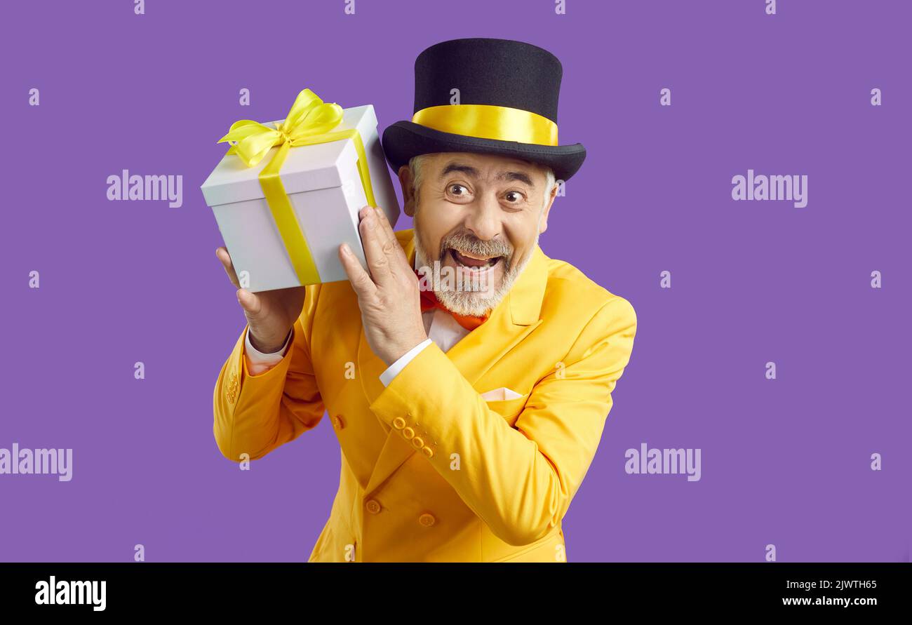 Gioioso uomo anziano divertente in abiti luminosi che tiene la scatola del regalo nelle sue mani su sfondo viola. Foto Stock