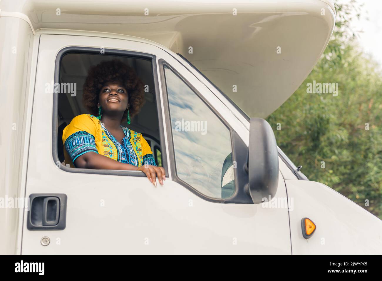 Giovane donna africana sorridente dalla pelle scura con taglio di capelli afro, indossando un abito boho luminoso, guardando fuori dalla finestra di un camper e ammirando la natura tranquilla intorno alla fine del suo viaggio. Foto di alta qualità Foto Stock
