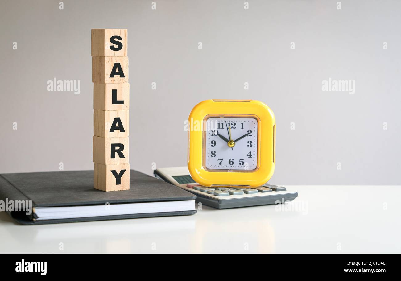 Testo dello stipendio sul cubo di legno, sulla parte superiore del blocco note e accanto alla calcolatrice e all'orologio. Foto Stock