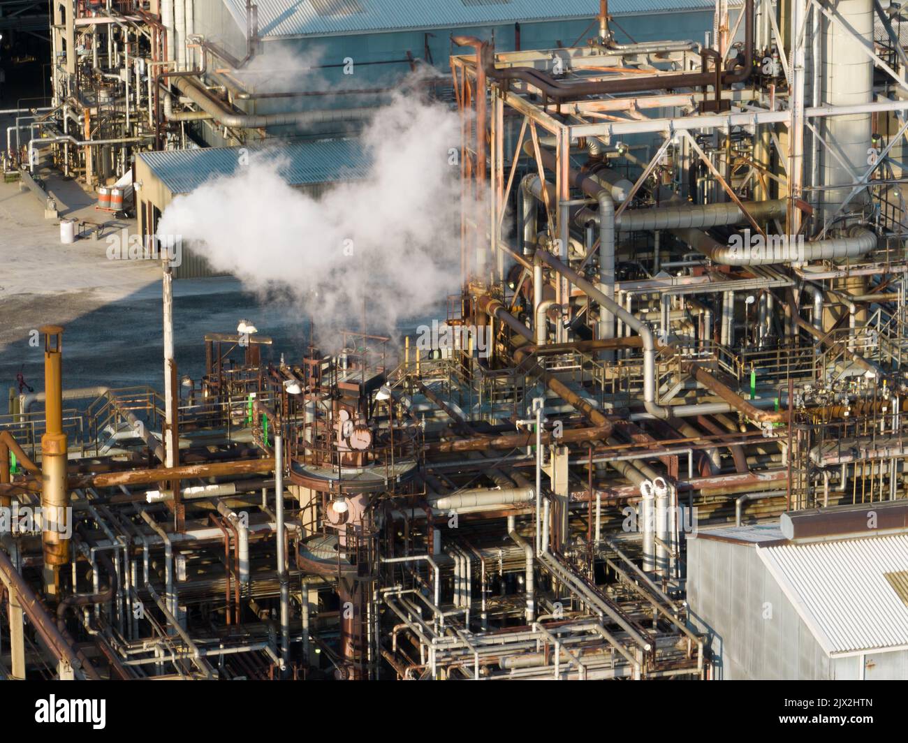 Una vista in primo piano di tubazioni industriali complesse in una raffineria di petrolio è vista durante la luce del mattino come un camino di scarico in funzione. Foto Stock