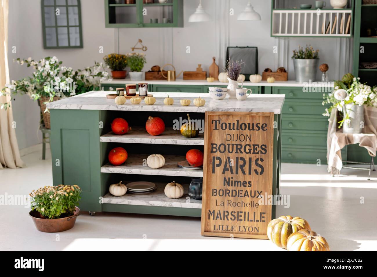 Cucina con mobili verdi vintage, lampade a sospensione, piano in marmo con fiori e zucche, pentole di fiori autunnali, armadio con varie tazze Foto Stock