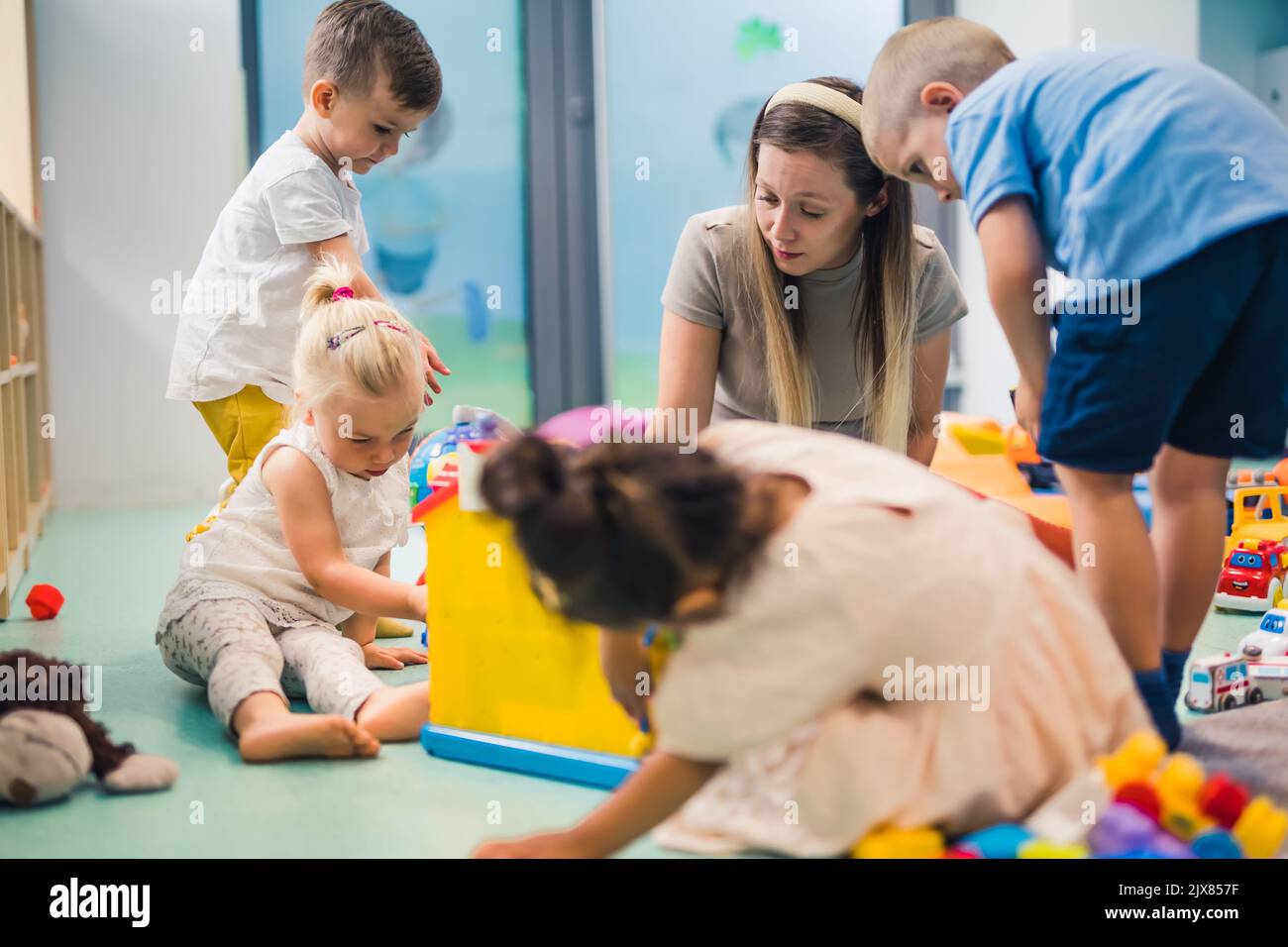 I bambini e il loro insegnante di asilo giocano con blocchi di plastica e giocattoli colorati per auto mentre si siedono sul pavimento in una sala giochi. Concentrazione, sviluppo delle capacità motorie fini e lorde, . Foto di alta qualità Foto Stock