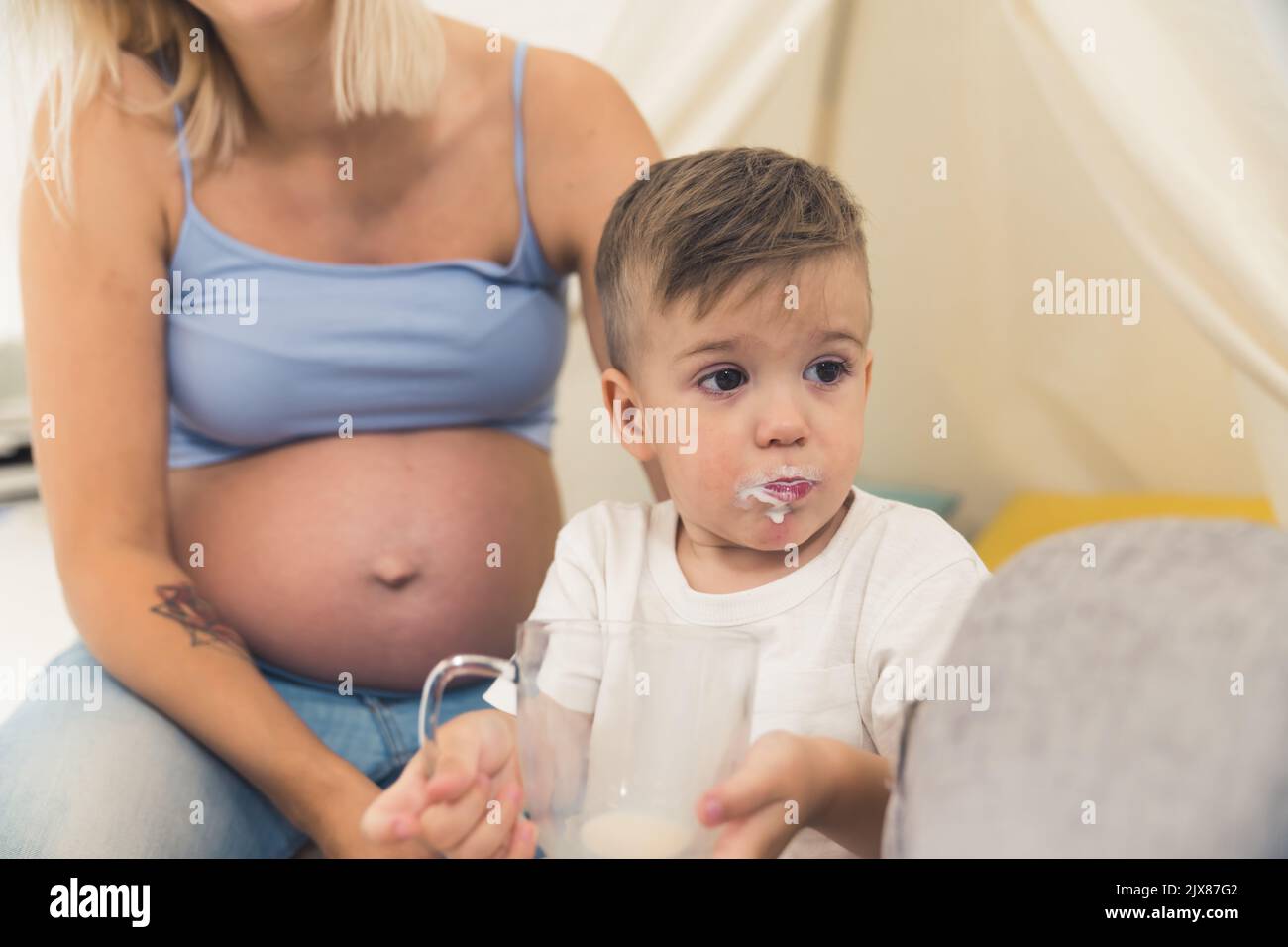 Divertente colpevole bambino caucasico bambino preschooler bere tutto il latte da una tazza. Il latte si marca intorno alle labbra. Madre incinta con pancia visibile sullo sfondo. Foto di alta qualità Foto Stock