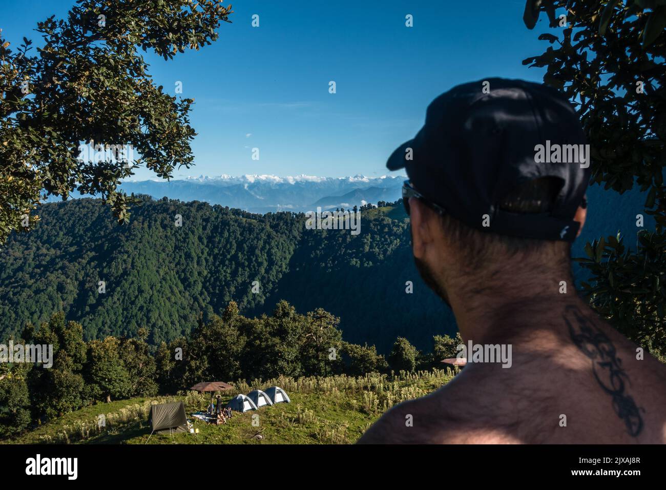 Settembre 16th 2021, Nag tibba montagna Uttarakhand India. Un uomo con un tatuaggio sulla schiena che guarda i paesaggi e le tende del campo base formano un'altezza dentro Foto Stock