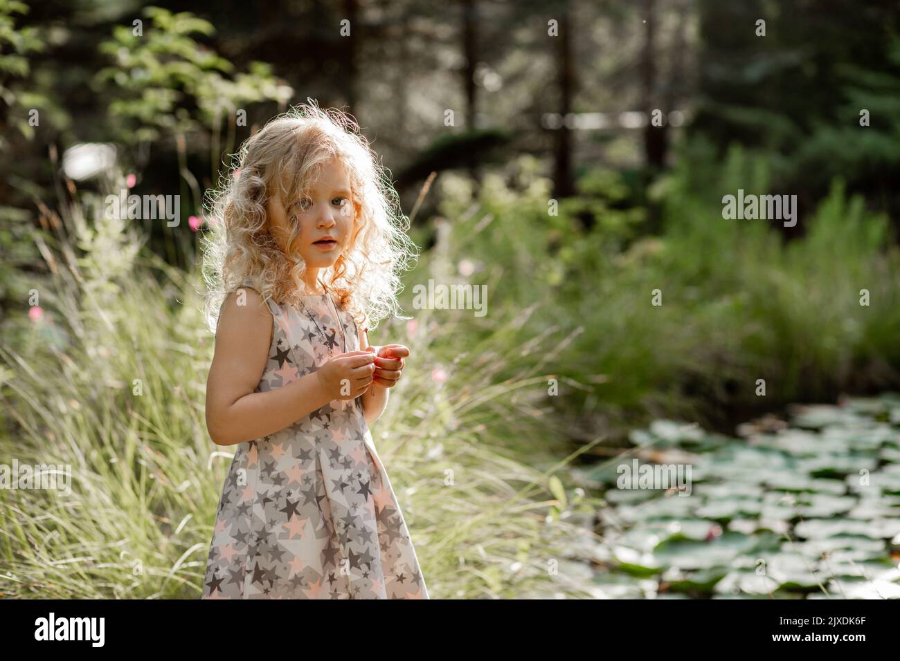 Ritratto di stravolgimento piangendo bambina con lunghi capelli ricci soffici in piedi vicino stagno con ninfee verdi nel parco. Foto Stock