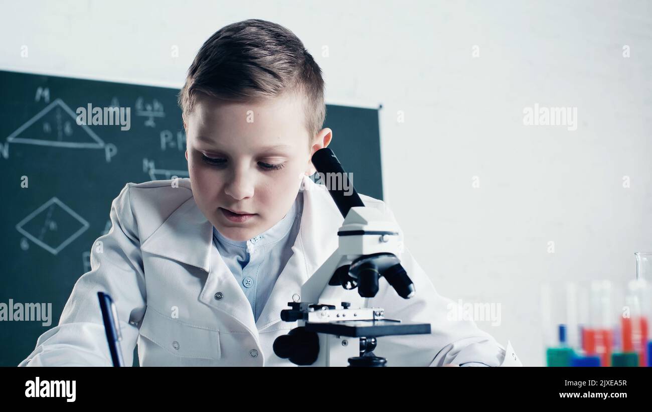 scolaro in camice bianco scrivere vicino al microscopio durante la lezione di chimica in classe, immagine stock Foto Stock