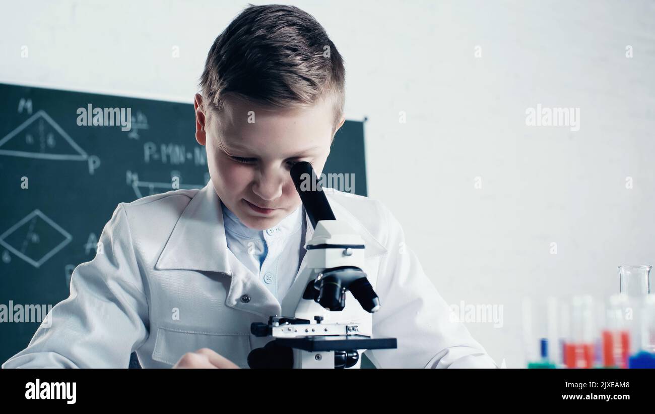 ragazzo in cappotto bianco che guarda attraverso il microscopio durante la lezione di chimica in classe, immagine stock Foto Stock