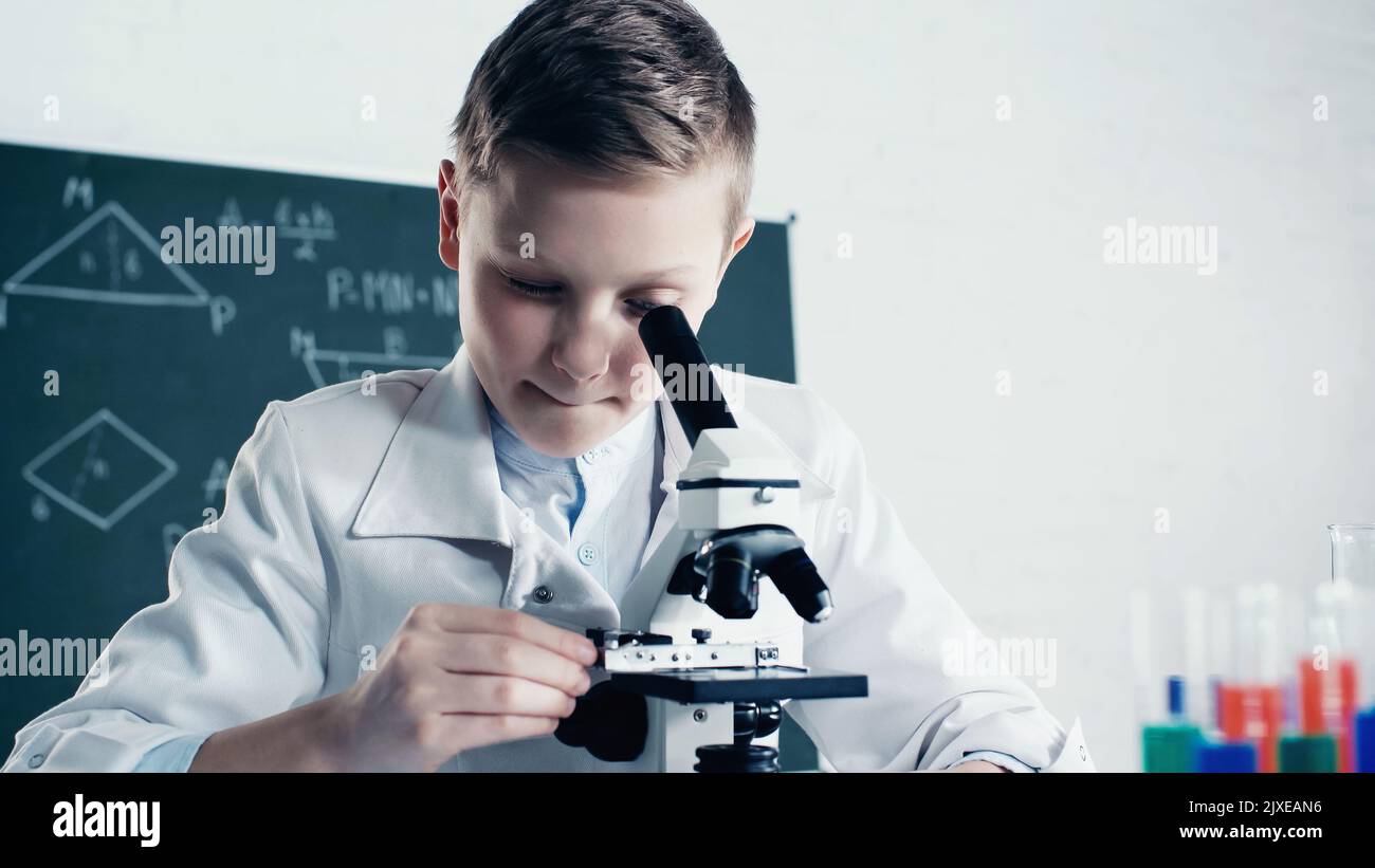 schoolkid in cappotto bianco guardando attraverso il microscopio durante la lezione di chimica in classe, immagine stock Foto Stock